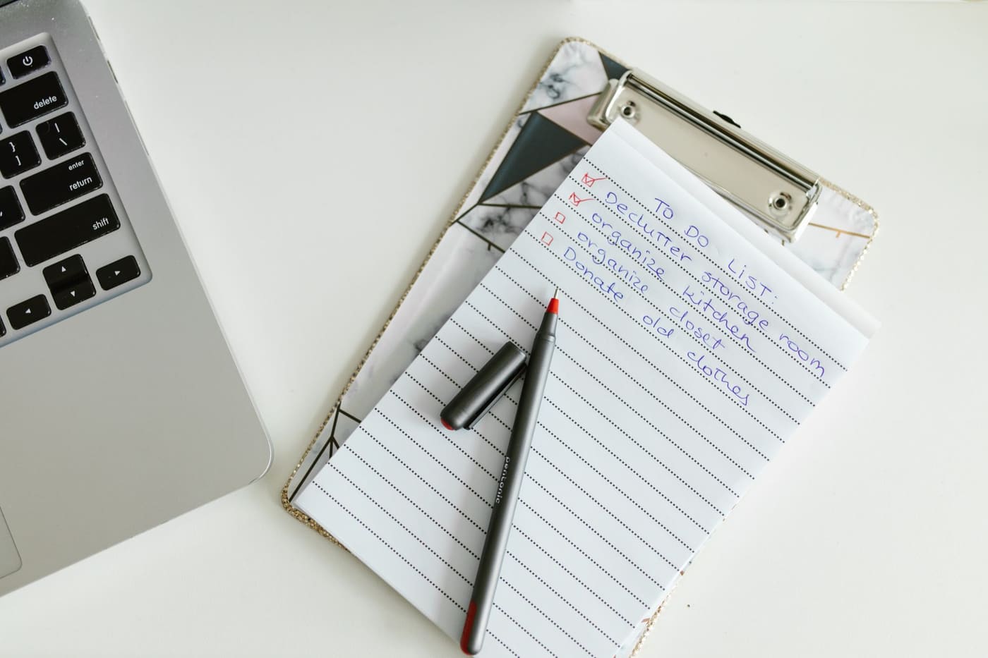 A lined notepad on a clipboard with a pen beside a laptop on a white desk