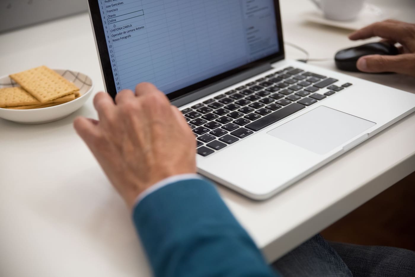 A hand on a laptop touchpad with a list on screen and a bowl of crackers nearby