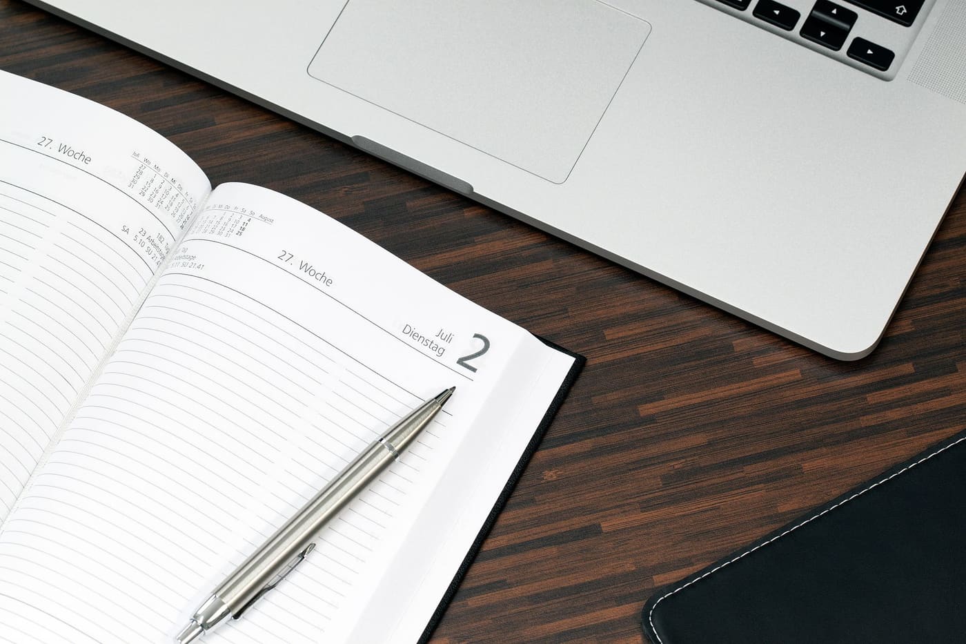 An open planner with a pen beside a laptop on a wooden desk