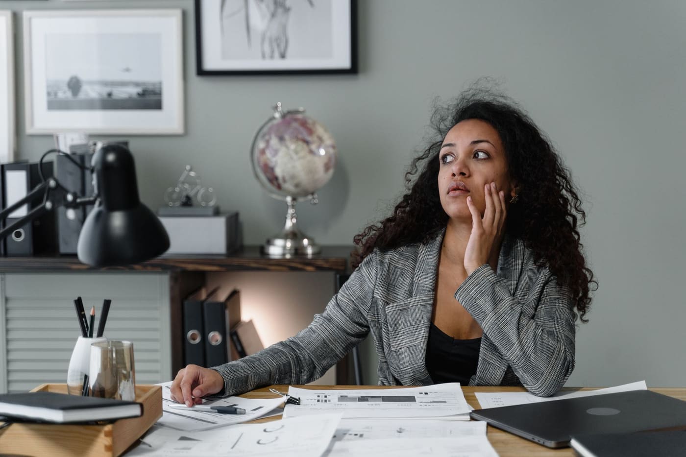 A woman in a plaid blazer thinking at her desk among papers and a laptop